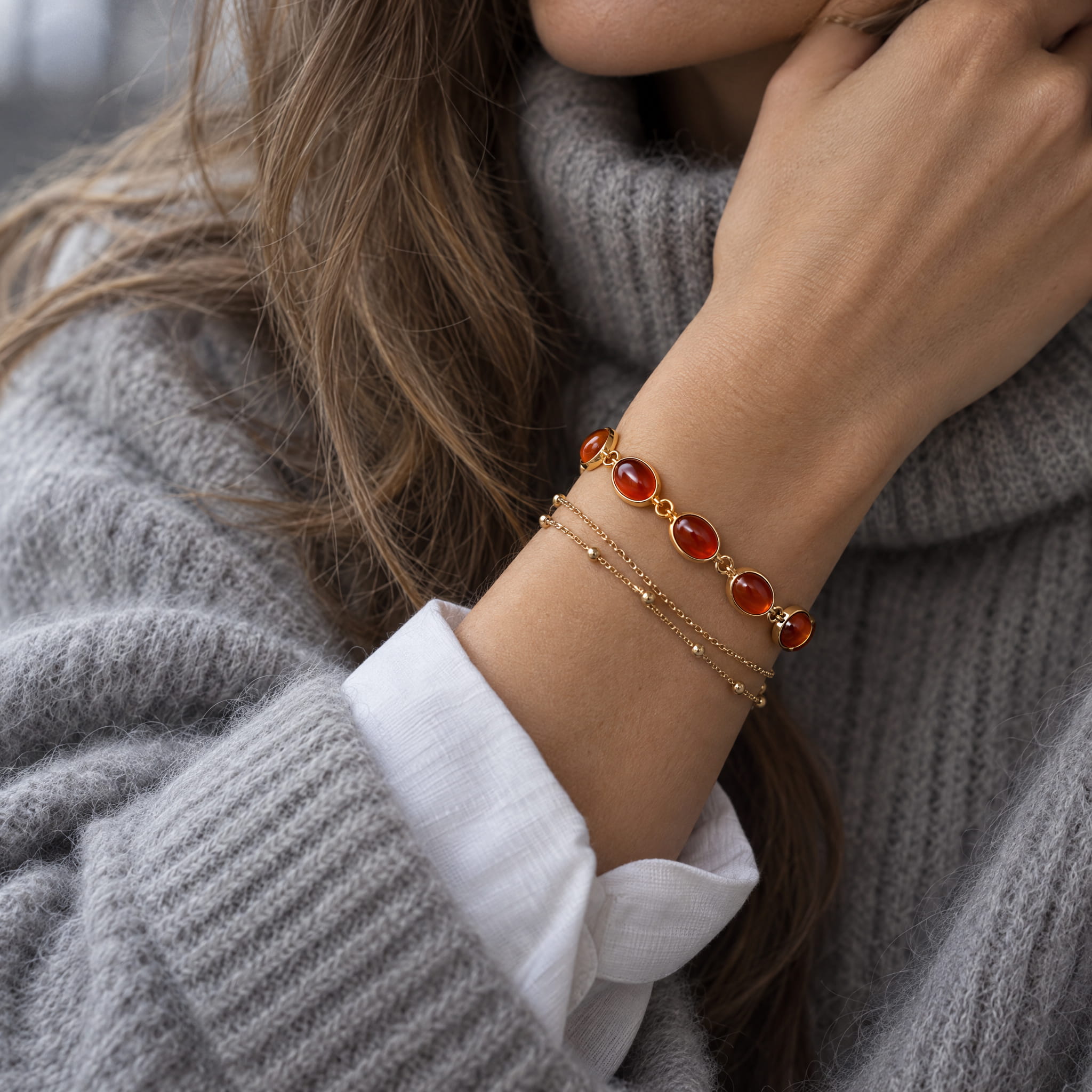 A close-up of a woman's wrist wearing two delicate gold bracelets, one featuring oval red-orange cabochon gemstones and the other a fine beaded chain, styled with a gray knit sweater and white collared shirt.
