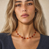 A close-up of a woman wearing a delicate gold chain necklace with polished amber-orange beads, set against a neutral beige background.