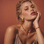 A woman wearing a matching amber-orange beaded necklace and bracelet with textured gold-toned hoop earrings, posed against a warm terracotta background.