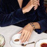 A close-up lifestyle shot of a person wearing a navy blue pinstripe velvet jacket and gold bracelets with blue stones, resting their hand on a white ceramic cup on a table covered in a white cloth.