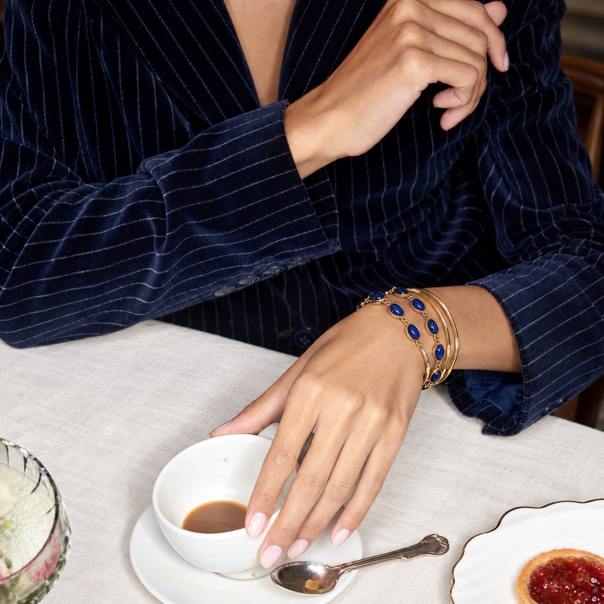 A close-up lifestyle shot of a person wearing a navy blue pinstripe velvet jacket and gold bracelets with blue stones, resting their hand on a white ceramic cup on a table covered in a white cloth.