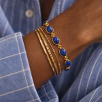 A close-up of a person's wrist wearing a stack of gold bracelets, including one with oval blue lapis lazuli stones, over a blue and white checkered shirt.