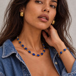 A woman wearing a gold and blue jewelry set, featuring a necklace, matching bracelet, and earrings, styled with an open denim shirt against a neutral gray background.
