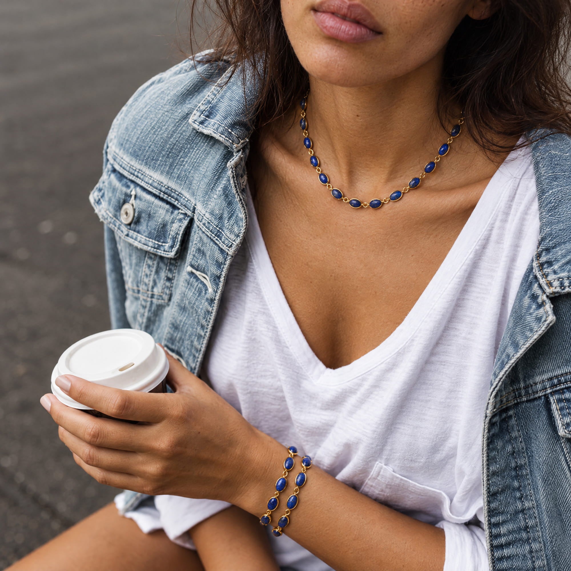 A close-up lifestyle shot of a woman wearing a matching beaded necklace and bracelet. The jewelry features deep blue oval beads set in gold-toned metal, styled with a white t-shirt and an open denim jacket while holding a takeaway coffee cup.
