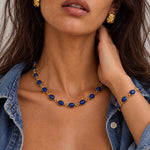 A close-up lifestyle shot of a model wearing a gold necklace and bracelet set with oval blue gemstones, paired with a denim shirt.