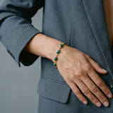 A gold chain bracelet with green oval stones worn on a person's wrist, shown against a gray blazer.