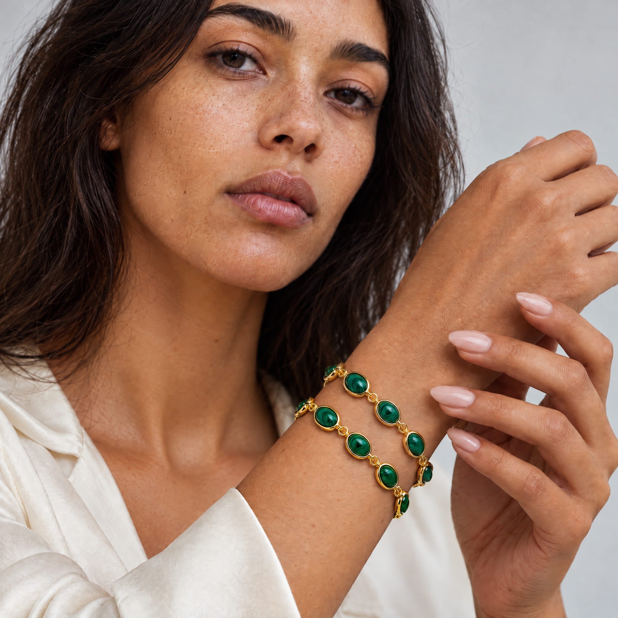 A close-up lifestyle shot of a woman wearing a double-strand gold bracelet with green oval cabochon stones, shown on her wrist against a soft gray background.