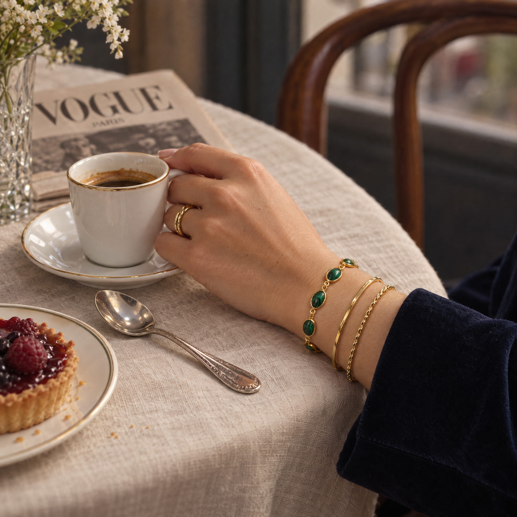 A close-up lifestyle shot of a person's hand resting on a cup of coffee at a cafe table, wearing a stack of gold bracelets, including one with green stones and two slim gold bangles. A VOGUE Paris newspaper, a glass, and a berry tart are visible on the table.