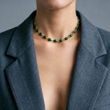 A close-up of a model wearing a gold chain necklace with green oval gemstones, styled with a dark gray blazer.