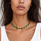 A close-up of a woman wearing a gold chain necklace with green oval gemstones, paired with a white ribbed tank top.