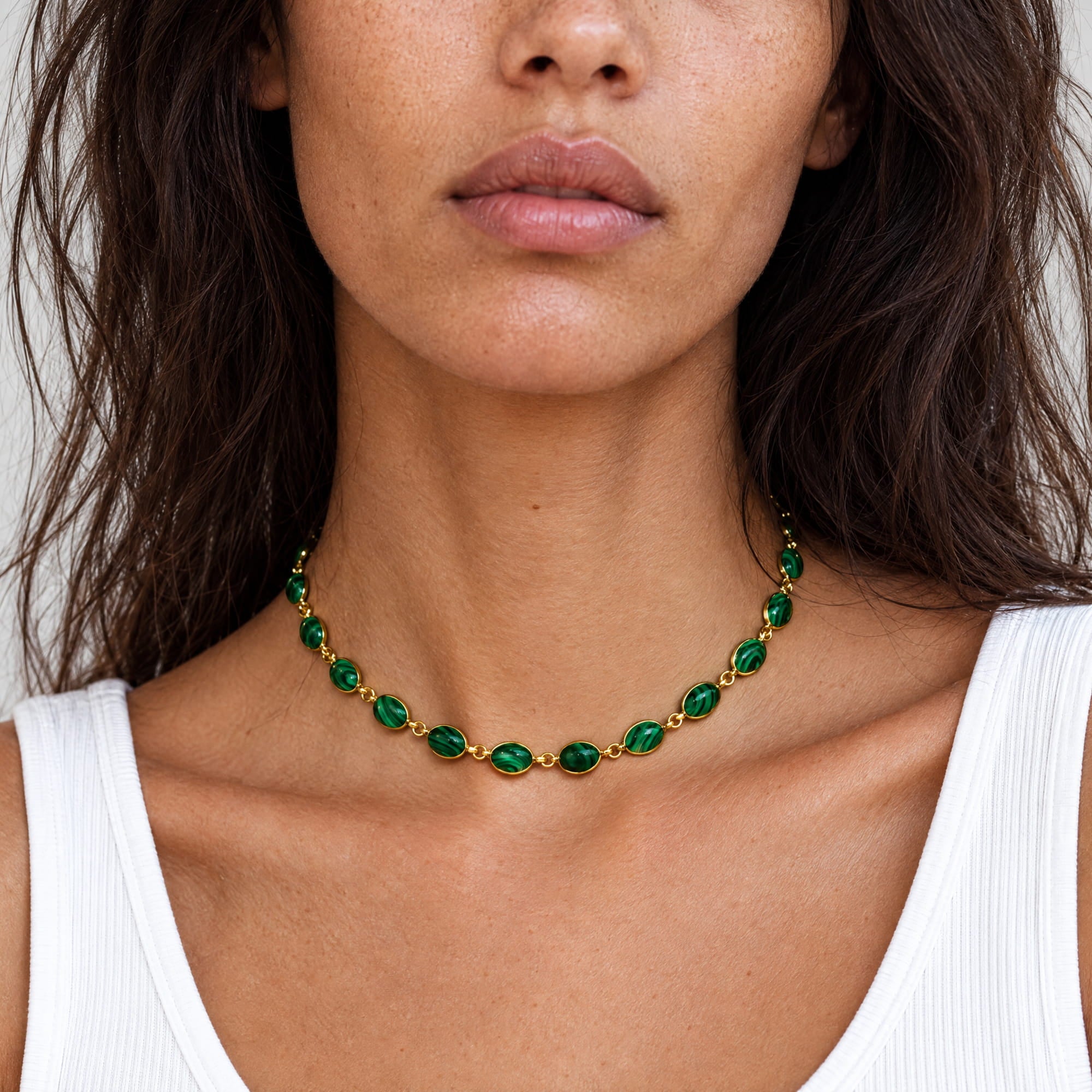 A close-up of a woman wearing a gold chain necklace with green oval gemstones, paired with a white ribbed tank top.
