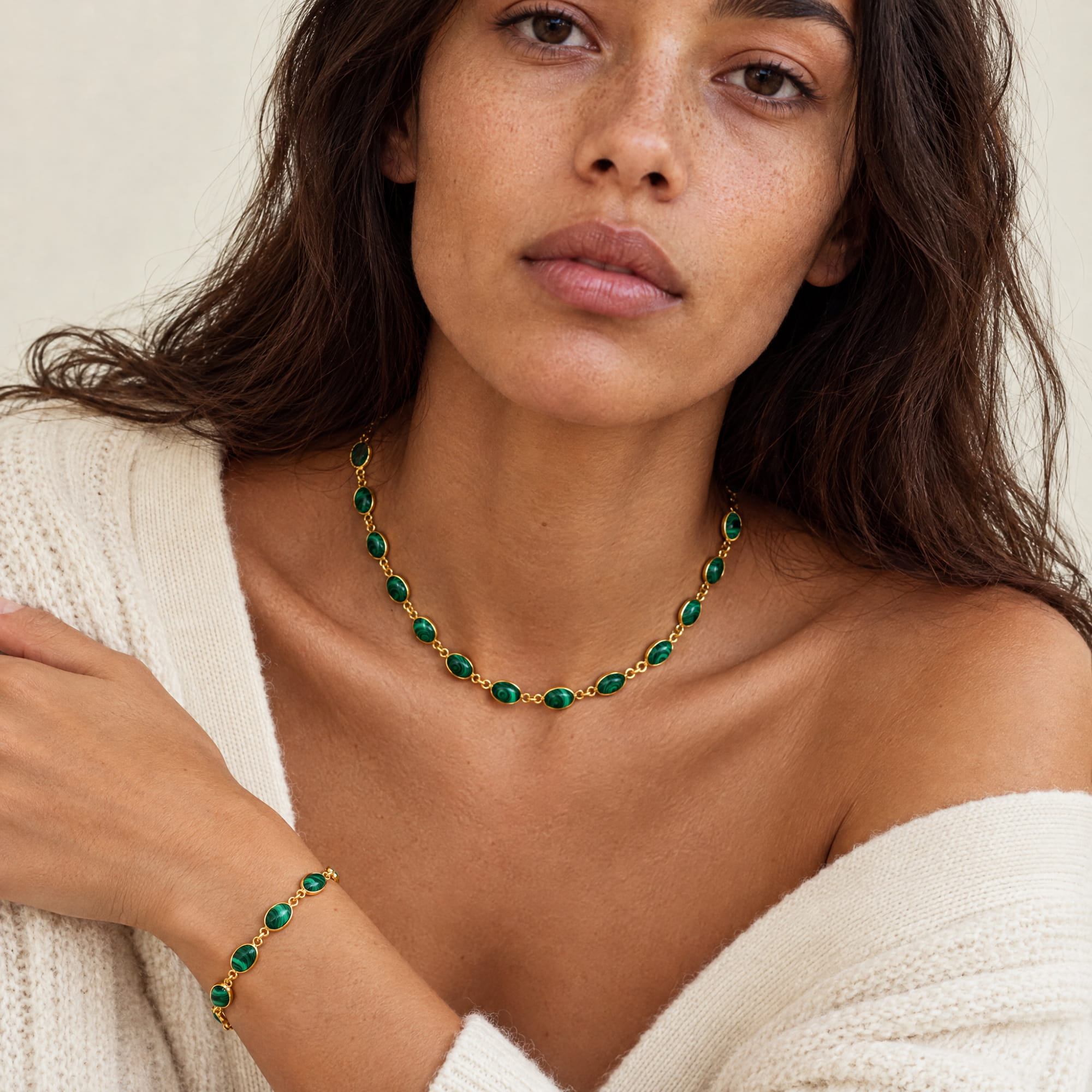 A close-up lifestyle shot of a woman wearing a gold necklace and matching bracelet with green oval stones, paired with a cream knit sweater.