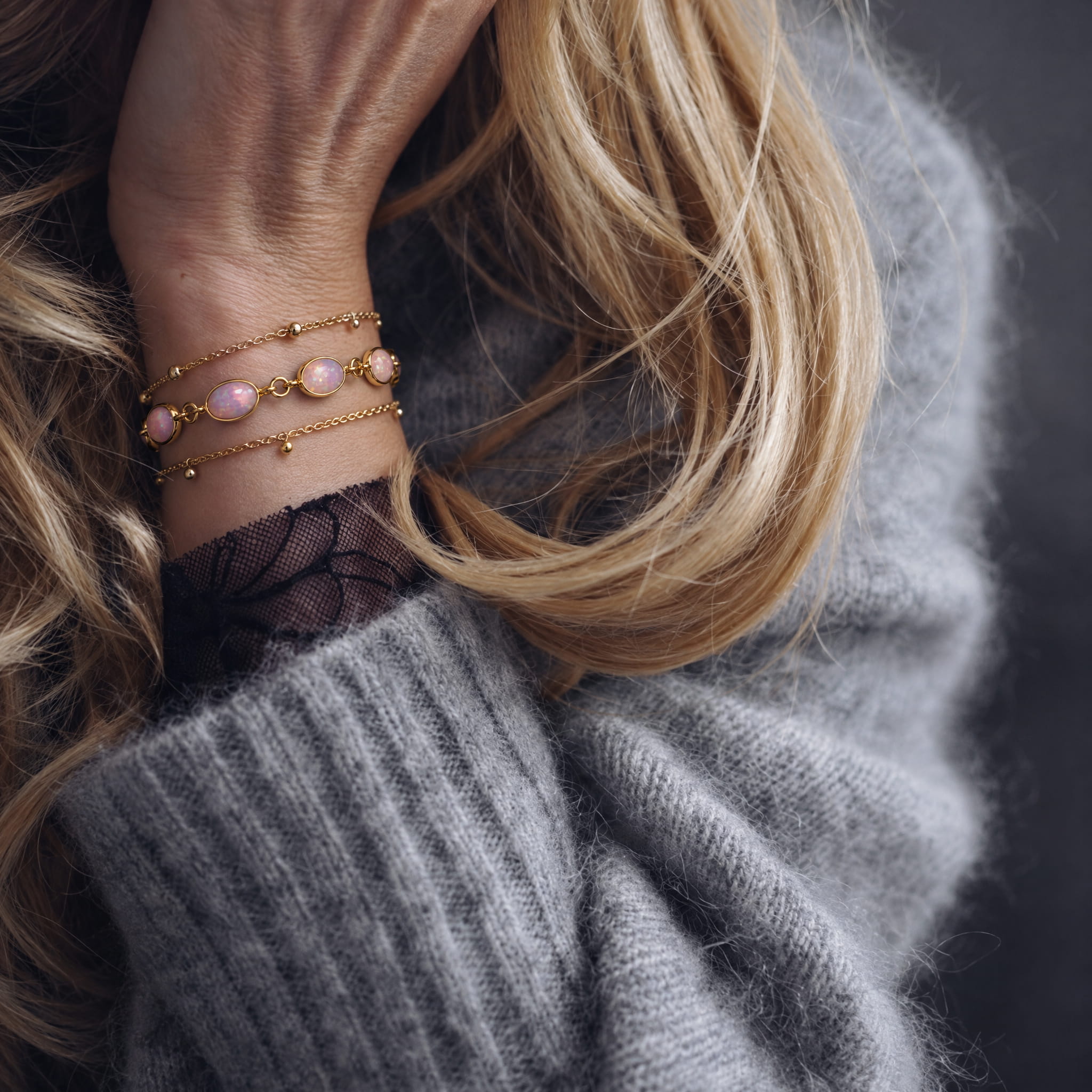 A close-up of a person's wrist wearing three delicate gold bracelets, including two with pale pink oval beads and a thin chain with small gold beads, paired with a gray knit sweater.
