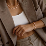 A close-up lifestyle shot of a person wearing a delicate pink beaded bracelet on their wrist and a matching pink beaded necklace on their neck, styled with a white ribbed tank top and a taupe blazer.
