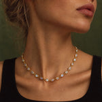 A close-up of a woman wearing a delicate gold chain necklace with oval iridescent white stones, set against a dark olive green background.