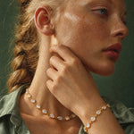 A close-up lifestyle shot of a woman with blonde hair in a braid, wearing a gold chain necklace and matching gold bracelet with white cabochon stones, set against a dark green background.