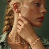 A close-up lifestyle shot of a woman with blonde hair in a braid, wearing a gold chain necklace and matching gold bracelet with white cabochon stones, set against a dark green background.