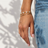 A close-up of a person's hand wearing two delicate gold bracelets, one with small white oval stones and one plain gold bangle, next to blue denim jeans against a white wall with leaf shadows.