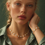 A close-up portrait of a woman wearing a gold chain necklace and matching bracelet with oval milky white stones, set against a dark green background.