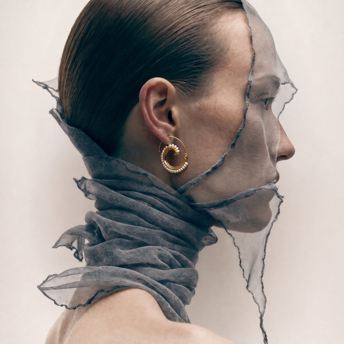 A close-up side profile of a model wearing a gold hoop earring with a textured beaded border, styled with a sheer gray scarf draped around the neck and face against a light beige background.