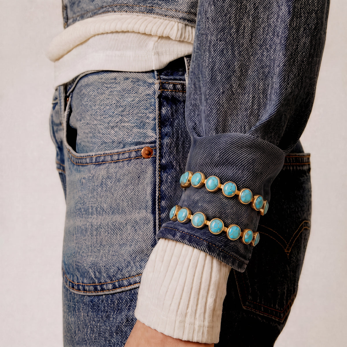 Close-up of a person wearing a dark blue denim jacket with a rolled cuff decorated with three rows of turquoise stones in gold settings, paired with blue jeans and a cream ribbed sweater.