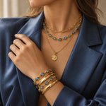 A close-up of a woman wearing layered gold jewelry, including a necklace with turquoise-blue stones and a stack of bracelets, paired with a navy blue blazer.