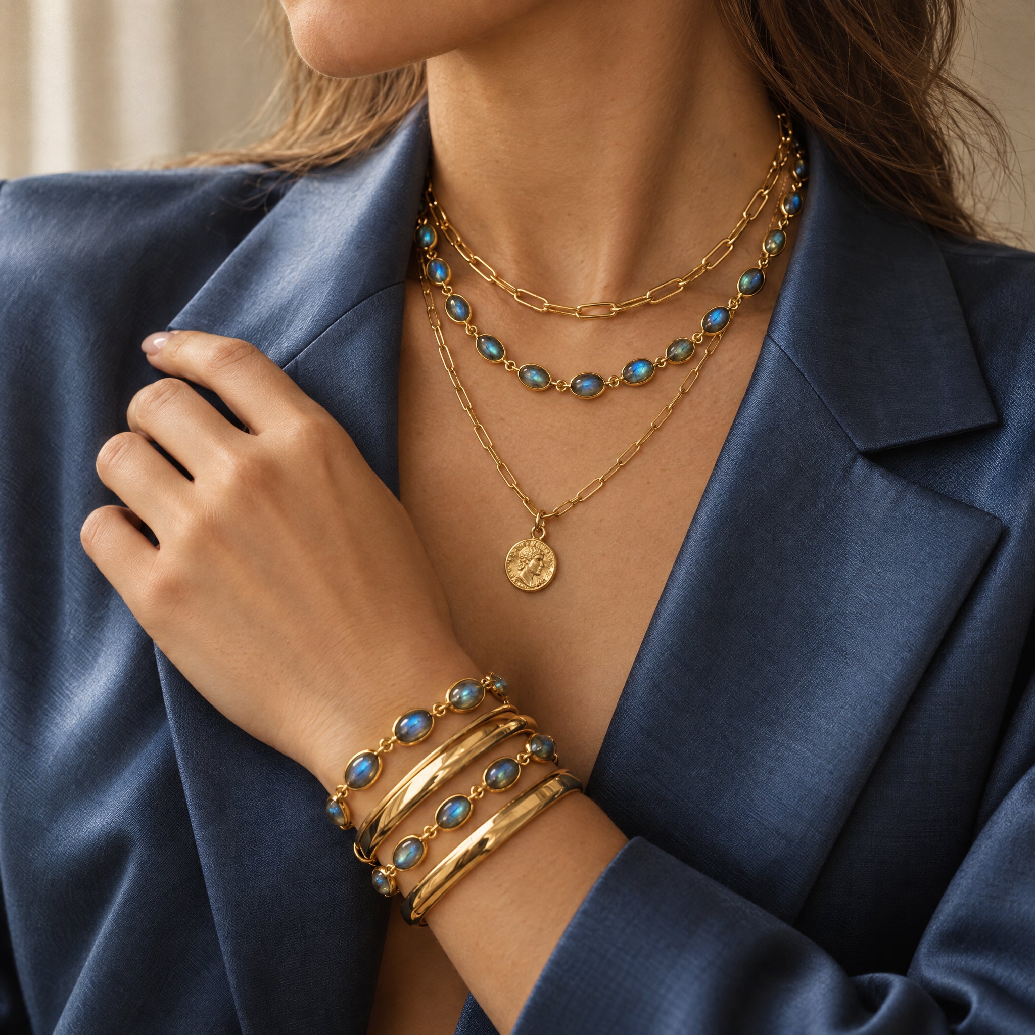A close-up of a woman wearing layered gold jewelry, including a necklace with turquoise-blue stones and a stack of bracelets, paired with a navy blue blazer.