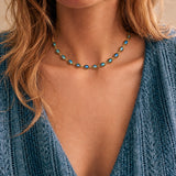 A close-up of a woman wearing a delicate gold chain necklace with small oval turquoise-blue beads, paired with a blue knit top.