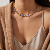 A close-up of a model wearing a double-layered gold necklace with oval blue-green iridescent stones, styled with a beige camisole and a brown cardigan.