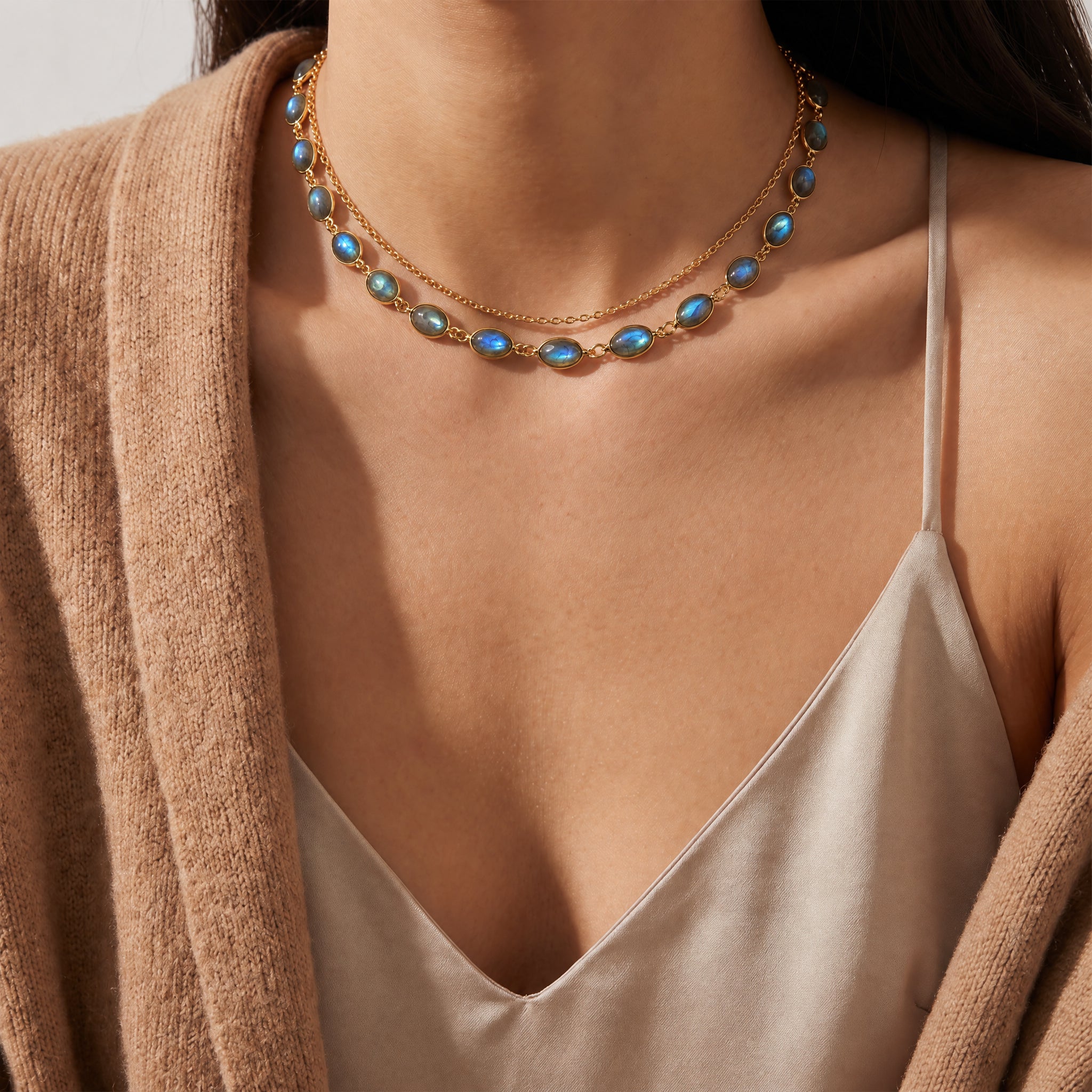 A close-up of a model wearing a double-layered gold necklace with oval blue-green iridescent stones, styled with a beige camisole and a brown cardigan.