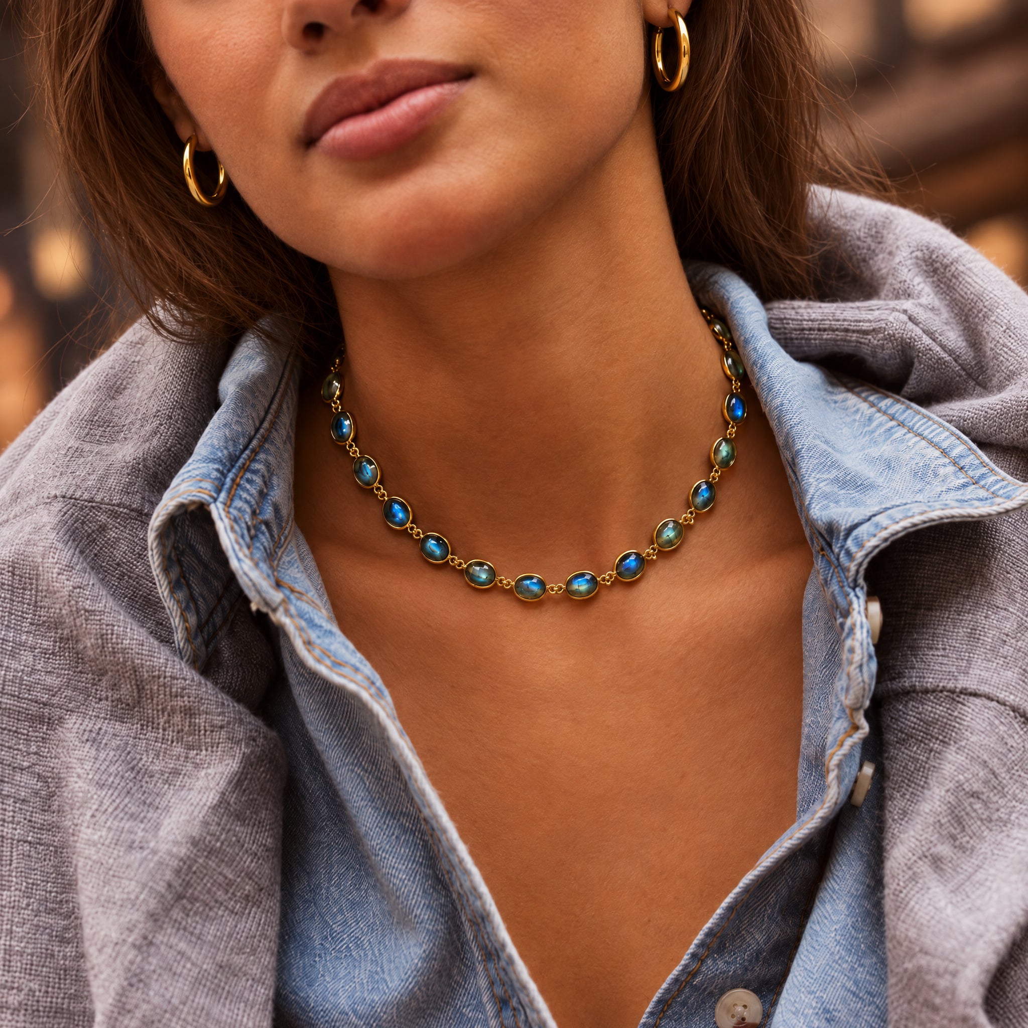 A close-up lifestyle shot of a woman wearing a gold chain necklace with oval blue-green stones, styled with gold hoop earrings and a light blue denim shirt.
