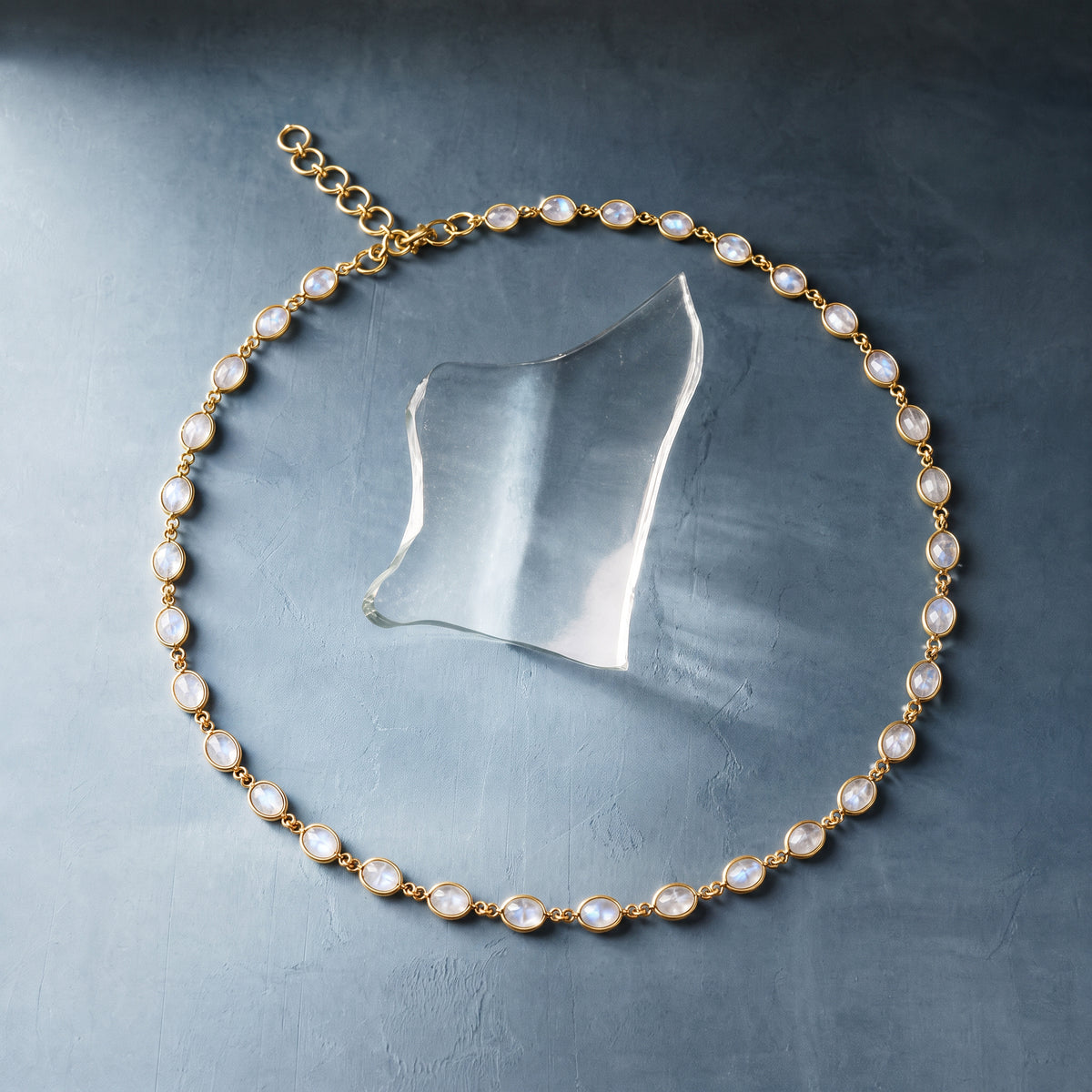 A gold chain necklace with oval white pearl-like stones, arranged in a circle on a textured dark gray surface with a clear glass prop.
